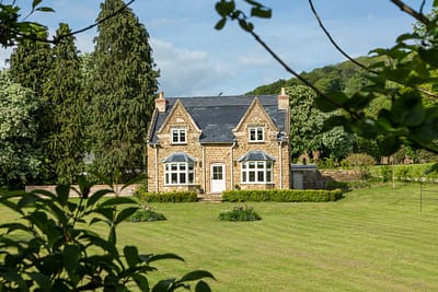 Underwood 1 Large A charming stone cottage with two chimneys, large windows, and a neatly trimmed lawn, surrounded by green trees and bushes on a sunny day. David Pearson Travel