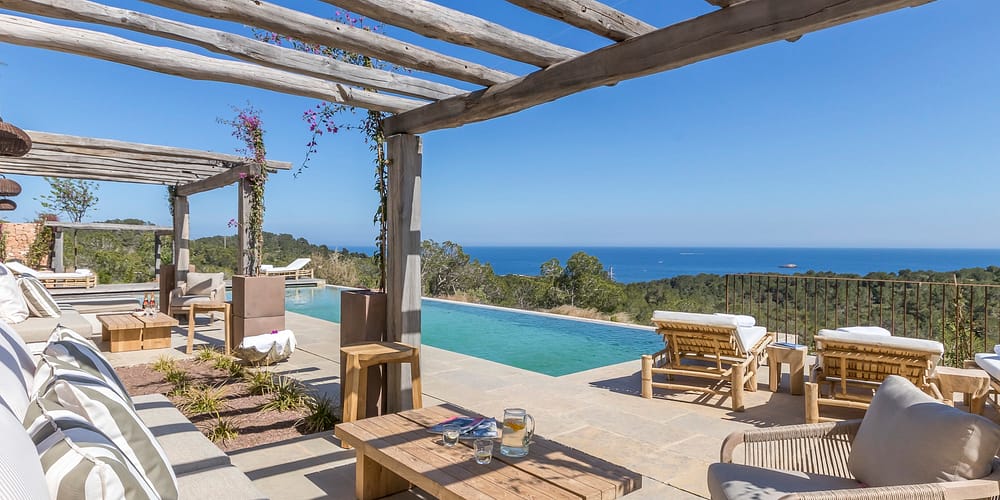 BBI-by Ali Framil-033 A luxurious outdoor patio with a pergola overlooks an infinity pool and a scenic ocean view, perfect for indulging in luxury travel. The area is furnished with comfortable seating and wooden lounge chairs. The sky is clear and blue, enhancing the serene atmosphere. David Pearson Travel