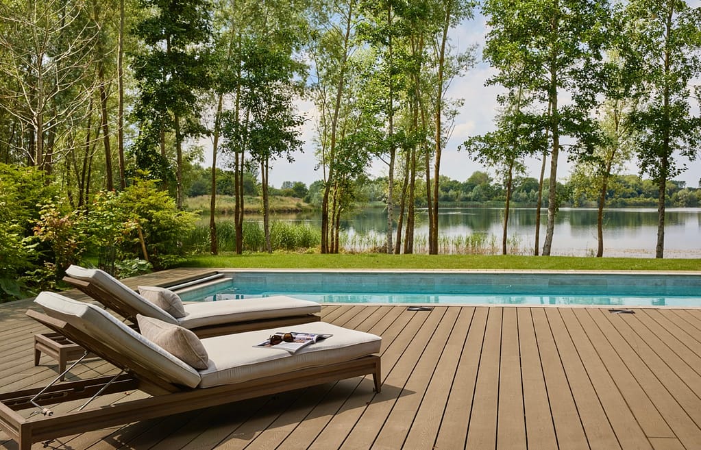 Swan Lake House - swimming pool side view Two cushioned lounge chairs sit on a wooden deck beside a swimming pool, overlooking a scenic lake surrounded by trees and greenery under a bright, partly cloudy sky. David Pearson Travel
