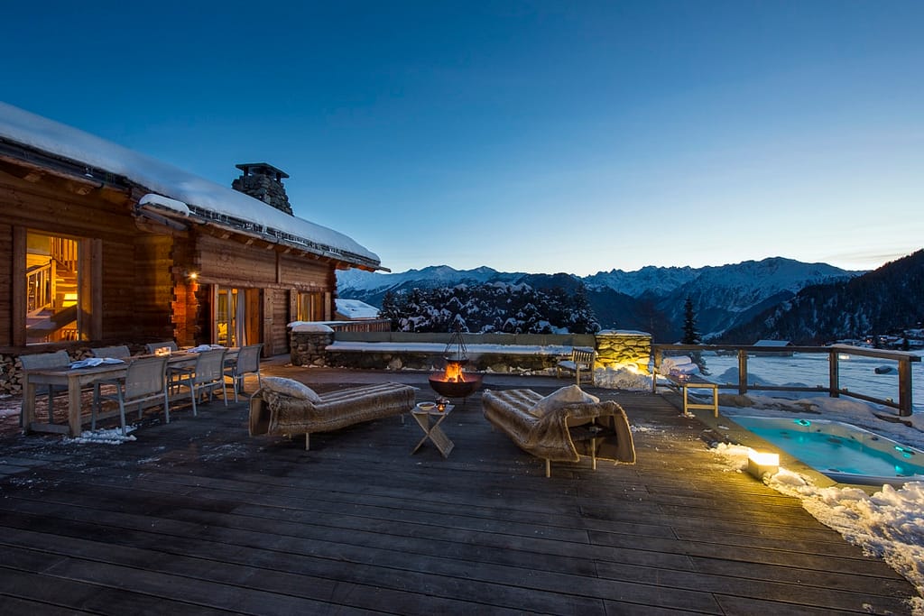 At dusk, the alpine cabin terrace offers a luxurious travel experience with its wooden deck, cozy seating, and inviting firepit. Snow blankets the area, while a hot tub with glowing lights adds warmth. The scene is completed by snow-capped mountains and a clear, darkening sky. David Pearson Travel