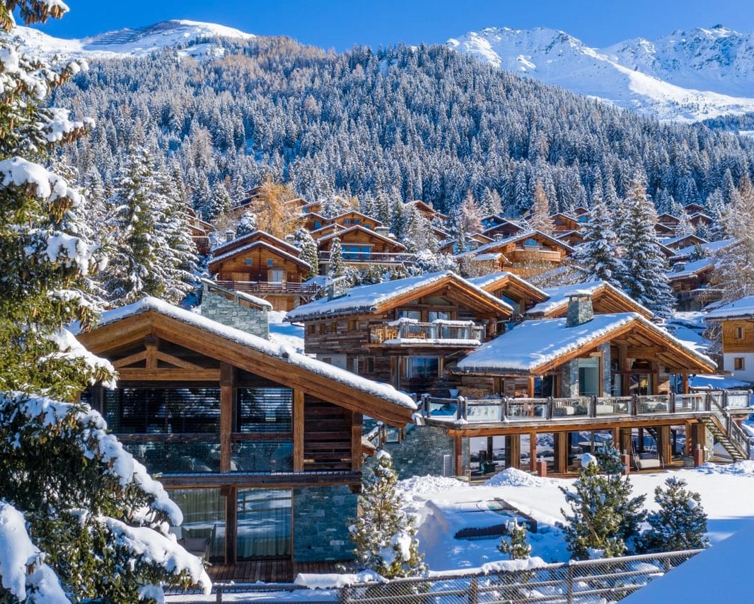 Verbier A cluster of wooden chalets with large windows sits in a snowy alpine village, perfect for your Home Page backdrop, surrounded by pine trees and mountains under a clear blue sky. Snow blankets rooftops and ground, creating a cozy wintery atmosphere. David Pearson Travel