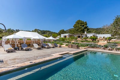 Ses Torres by Ali Framil-03 Experience the essence of luxury travel in this exquisite outdoor setting, where a clear swimming pool glistens alongside lounge chairs and white canopy tents on a wooden deck. Behind lies an elegant house nestled amid lush greenery and trees, all under a bright blue sky. David Pearson Travel