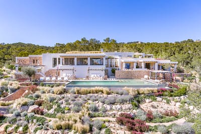 BBI-by Ali Framil-149 A luxurious hilltop house with a modern design, perfect for luxury travel enthusiasts, is surrounded by lush greenery and gardens. The property features a large rectangular swimming pool and expansive outdoor seating areas, set against a clear blue sky backdrop. David Pearson Travel