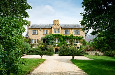 Exterior down drive A large, picturesque stone house with ivy-covered walls stands at the end of a gravel path, offering a luxury travel experience. Surrounded by lush greenery and trees, the two-story building has multiple windows and a chimney, set under a partially cloudy sky. David Pearson Travel