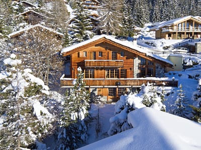 Les-Attelas-Exterior-1-scaled A cozy wooden chalet, ideal for luxury travel, is surrounded by snow-covered trees and hills in a winter landscape. With a stone foundation, large windows, and balconies, it stands amidst other similar houses. Snow blankets the roof and surroundings, creating a serene escape. David Pearson Travel