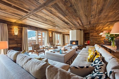 PlaceBlanche1 - Living Room (13) Spacious living room with wooden walls and ceiling, featuring a luxury travel-inspired large sectional sofa with colorful cushions, a central coffee table, and a fireplace. Large windows offer a view of mountains, and natural light fills the room. David Pearson Travel