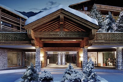 Rosalp3 - Exterior Rue de Medran (1) A luxurious mountain hotel entrance, epitomizing luxury travel, features a wooden and stone facade adorned with snow. The building glows warmly beneath a snowy blanket, encircled by snow-laden pine trees, crafting a cozy winter retreat. David Pearson Travel