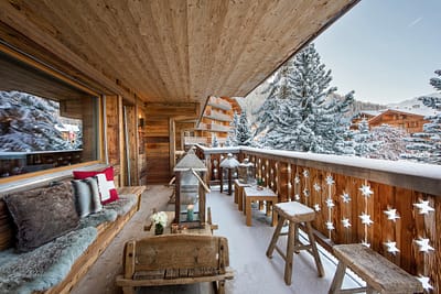 Rosalp4 - Balcony (2) A cozy wooden balcony with rustic seating offers a luxury travel experience, overlooking a snowy landscape with evergreen trees and wooden chalets. The railing, adorned with star cut-outs, complements the fluffy cushions and blankets arranged on the bench. David Pearson Travel
