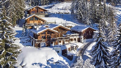 Slider_image_-_rsz_dji_0248_medium A luxury travel experience awaits at this snow-covered chalet nestled among pine trees on a hillside. The wooden building features multiple sloped roofs and balconies. A narrow path winds its way to the chalet, while the surrounding landscape is blanketed in snow under a clear blue sky. David Pearson Travel