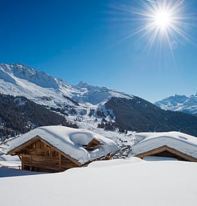 Nyumba - Exterior Rear (6) 3 Luxury travel takes center stage with snow-covered wooden cabins nestled in a mountainous landscape under a clear blue sky. The sun shines brightly, illuminating the white snow and surrounding pine trees, while majestic snowy peaks rise in the background, offering an unparalleled winter escape. David Pearson Travel