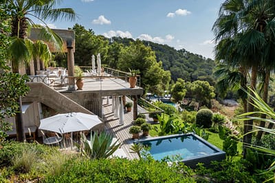 can-jacq-luxury-villa-ibiza-46-w1800-h950 A luxurious villa with a large terrace, outdoor dining area, and a small pool surrounded by lush greenery and palm trees, overlooking a scenic forested hillside under a partly cloudy sky. David Pearson Travel