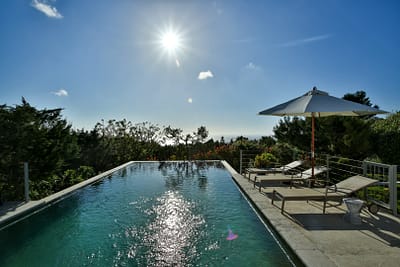 luxury-ibiza-calatarida-sanjose-25 A serene infinity pool reflects the bright sun with lounge chairs and an umbrella on the right, surrounded by lush greenery under a clear blue sky. David Pearson Travel