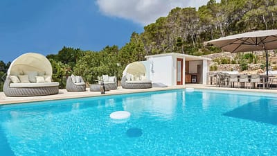 vanessa-12-w1850-h950 Luxurious outdoor pool with clear blue water, surrounded by modern round lounge chairs, umbrellas, and a small white poolside building, set against a backdrop of trees and blue sky. David Pearson Travel