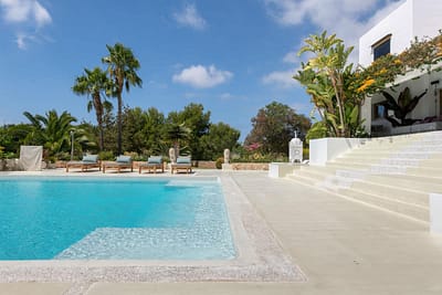 villa-lampy-luxury-villa-ibiza-14 A luxurious outdoor swimming pool with sun loungers, palm trees, and lush greenery around. White steps lead up to a modern white villa under a bright blue sky with scattered clouds. David Pearson Travel