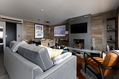 Haute'Xposure A cozy living room with a gray sofa, brown leather chair, wooden coffee table, and modern fireplace evokes the inviting charm found in luxury Verbier ski chalet rentals—art, blankets, and rich decor create a warm, welcoming atmosphere. David Pearson Travel
