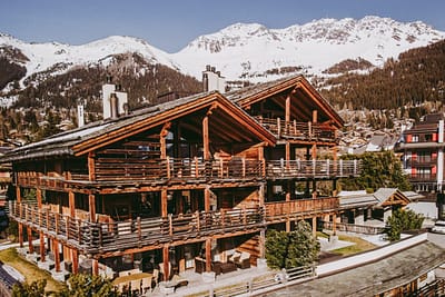 Ski_Armadillo_Verbier_Chalet_Vantine211-7 Large Large wooden chalet with balconies, set against snow-capped mountains and pine trees under a clear sky, surrounded by other alpine buildings. David Pearson Travel