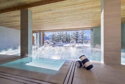 heated-swimming-pool-at-chalet-teredo A modern indoor pool with steam rising from the water, next to a large window overlooking snowy mountains and pine trees. Two rolled towels with blue and white stripes rest on the tiled floor. David Pearson Travel