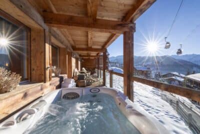 hot-tub-views-from-the-balcony-at-apartment-basalte A wooden chalet balcony features a bubbling hot tub overlooking snowy mountains and ski gondolas under a bright blue sky with sunlight streaming in. Cozy seating and rustic decor complete the inviting winter scene. David Pearson Travel