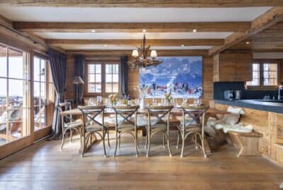 the-dining-table-at-apartment-silver A rustic dining room with a long wooden table, ten chairs, sheepskin bench, and a mountain scene photo on the wall. Large windows let in natural light; wood beams and floors create a cozy chalet ambiance. David Pearson Travel