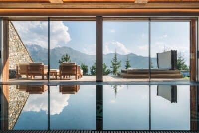 calima-ve-17 A luxurious indoor pool faces large glass doors opening to a terrace with wooden lounge chairs and a hot tub, overlooking mountains and pine trees under a clear sky. David Pearson Travel