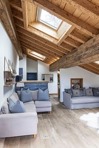 20250905-1120-4443-MSKY A cozy loft living room with wooden beams, light wood floors, a skylight, two gray sofas with blue and gray cushions, and a small open kitchen area in the background. David Pearson Travel