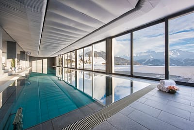 DSC_4240 An indoor pool with large floor-to-ceiling windows overlooking snowy mountains. Rolled towels and flowers are on the tiled floor beside the pool, and sunlight streams in, creating reflections on the water. David Pearson Travel