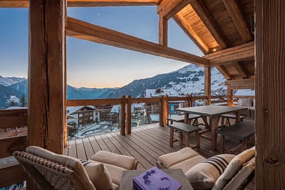 Falcon Penthouse 59 Cozy wooden balcony with lounge chairs, a table, and benches overlooking a snowy mountain village at sunset, with warm indoor lighting and scenic alpine views in the background. David Pearson Travel