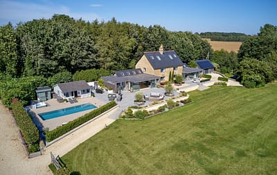 3C07BAF2-DBD1-4A74-9C2C-023087655286 Aerial view of a large countryside house with a stone exterior, surrounded by trees and fields. The property features a swimming pool, patio seating areas, outbuildings, landscaped gardens, and an expansive lawn. David Pearson Travel