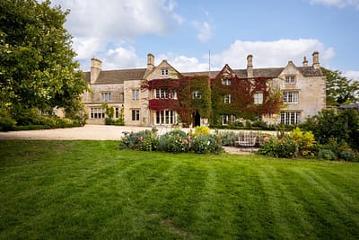 2. Exterior House Large Large stone manor house with ivy growing on its central walls, tall chimneys, many windows, surrounded by landscaped gardens and a green lawn under a partly cloudy sky. David Pearson Travel
