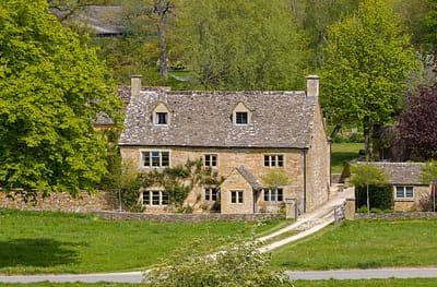 HL. 1 A traditional stone cottage with a steep roof, surrounded by lush green trees and grass, sits at the end of a gravel path in a rural countryside setting. David Pearson Travel