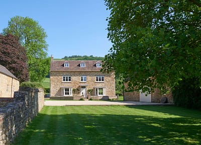 Farmhouse 1 A stone country house with a red roof sits under a clear blue sky, surrounded by green trees and a neatly mown lawn. A stone wall borders the left side of the scene. David Pearson Travel