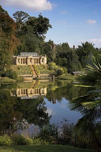 Temple 1 Large A small, elegant building with columns is reflected in a calm lake, surrounded by lush greenery and trees under a blue sky with clouds. David Pearson Travel