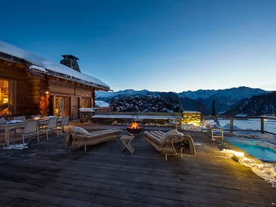 Pierre Avoi - Terrace (1) At dusk, the alpine cabin terrace offers a luxurious travel experience with its wooden deck, cozy seating, and inviting firepit. Snow blankets the area, while a hot tub with glowing lights adds warmth. The scene is completed by snow-capped mountains and a clear, darkening sky. David Pearson Travel