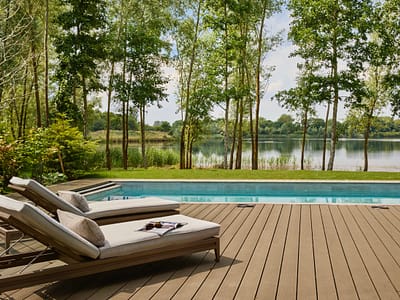 Swan Lake House - swimming pool side view Two cushioned lounge chairs sit on a wooden deck beside a swimming pool, overlooking a scenic lake surrounded by trees and greenery under a bright, partly cloudy sky. David Pearson Travel