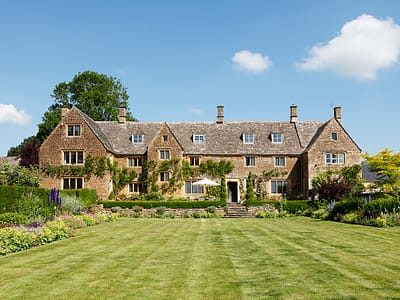 1 Large stone country house with steep roofs and multiple chimneys, surrounded by lush gardens and a neatly mown lawn, under a bright blue sky with a few clouds. David Pearson Travel