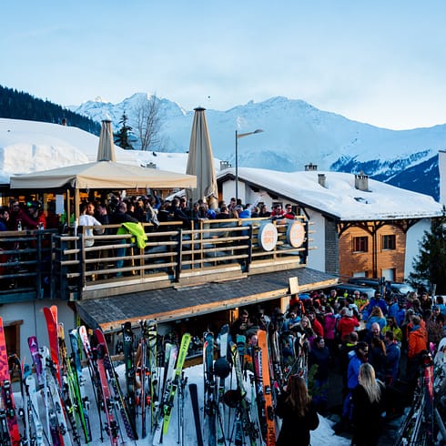 le roughe Crowds gather for après-ski at a lively outdoor alpine bar in Verbier, with snow-covered rooftops, colorful skis lined up outside, and snowy mountains in the background under a clear evening sky—perfect for Verbier travel enthusiasts. David Pearson Travel