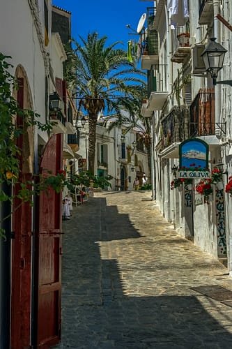 photo-1574516886768-cba7fe38ef97 A narrow, sunlit cobblestone street winds uphill between whitewashed buildings with balconies, potted plants, and palm trees. A colorful La Oliva sign hints at the charm of Ibiza travel. The sky is clear and blue. David Pearson Travel