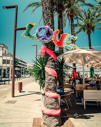 photo-1580381728525-8ec1964135e9 A palm tree on a sunny Ibiza street is decorated with colorful letters spelling LOVE and a peace sign, with pink fabric wrapped around the trunk. People sit at outdoor tables under umbrellas nearby. David Pearson Travel