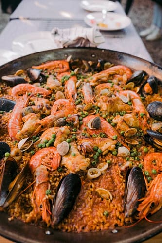 photo-1650964807311-970cb88d347c A large pan of seafood paella with mussels, shrimp, clams, peas, and rice, set on a table with plates and utensils in the background—perfect for savoring during your Ibiza travel experience. David Pearson Travel