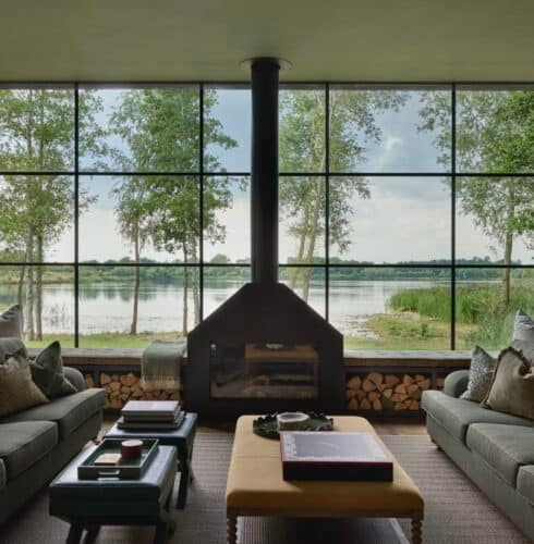 Swan Lake House - sitting room view A modern living room with two gray sofas, a central fireplace, large windows revealing a lake and trees outside, and neatly stacked firewood under the windows. Pillows and books decorate the space. David Pearson Travel