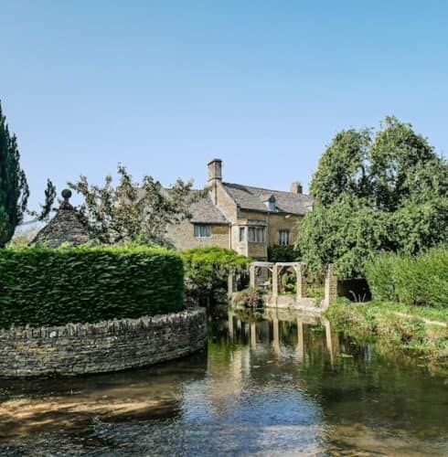 photo-1608221606535-15c75e4afb3c A stone cottage with a pitched roof sits beside a clear, shallow stream in The Cotswolds, surrounded by lush green trees and hedges under a bright blue sky. David Pearson Travel
