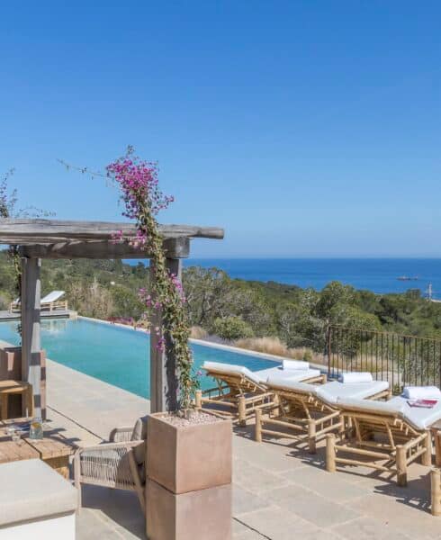 BBI-by Ali Framil-032 A scenic outdoor pool area with cushioned lounge chairs overlooks a vast, clear blue ocean, epitomizing luxury travel. The pool is bordered by a wooden pergola adorned with flowering vines, while lush greenery surrounds the area under a clear blue sky. David Pearson Travel