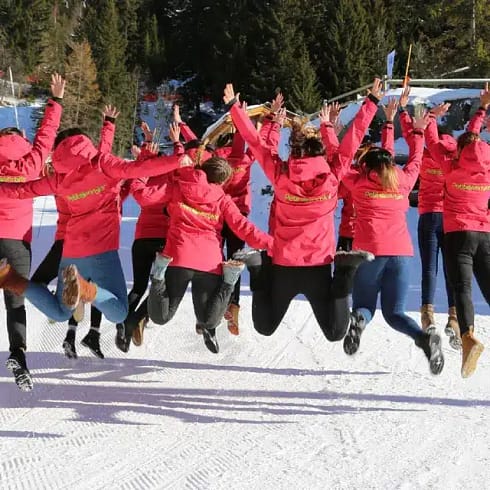 Petit Verbier A group of people in matching red jackets jump in the air on a snowy slope in Verbier, facing away from the camera, with trees and blue sky in the background—a perfect moment for Verbier tourism. David Pearson Travel