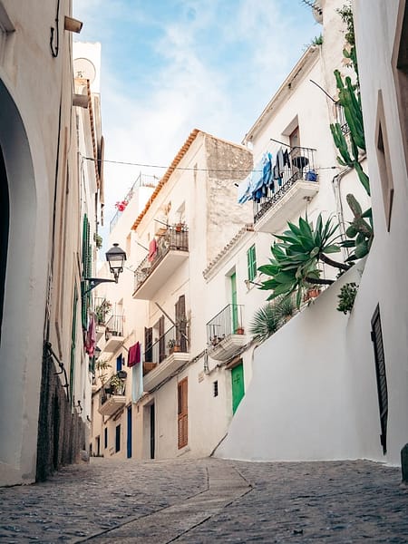 photo-1656081531767-a8d25cc06335 A narrow cobblestone street lined with whitewashed buildings, balconies displaying potted plants and colorful laundry, under a blue sky—an idyllic scene straight from an Ibiza guide for your next Ibiza travel adventure. David Pearson Travel