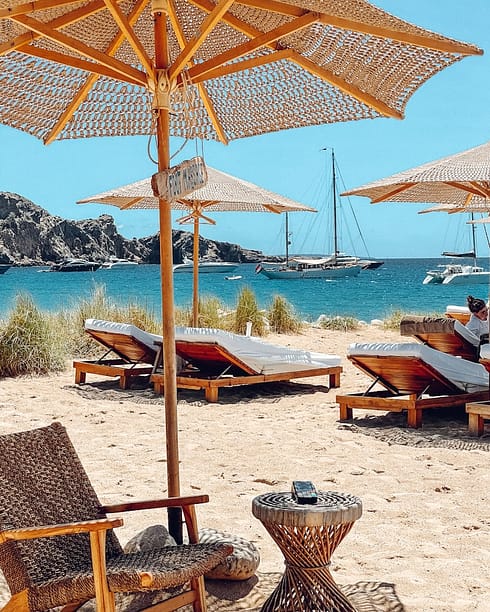photo-1622890806166-111d7f6c7c97 Beach scene with wooden lounge chairs and thatched umbrellas on sand, overlooking Ibiza’s blue sea with sailboats in the distance. Grassy dunes and a rocky hill are visible in the background under a clear sky. David Pearson Travel