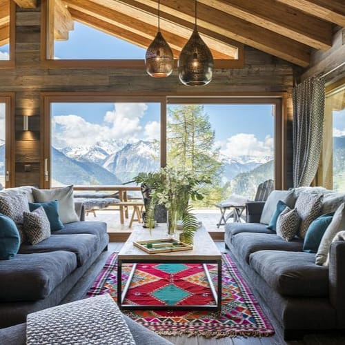 Chalet Mowgli David Pearson Travel A cozy living room perfect for your Home Page, with large windows revealing snowy mountains, two gray sofas, a colorful rug, and a wooden coffee table with flowers. Natural light highlights the wood beams and decorative pillows. David Pearson Travel