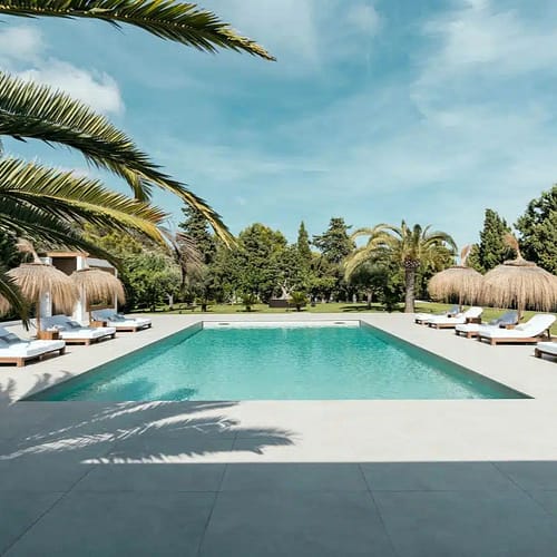 villa-maggie-22.jpg - Edited A rectangular outdoor pool surrounded by white lounge chairs and straw parasols, featured on the Home Page, with palm trees and greenery in the background under a blue sky. David Pearson Travel
